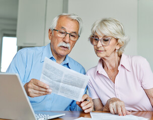 Senior couple checking bills and calculating expenses using a laptop at home, debt taxes concept,...
