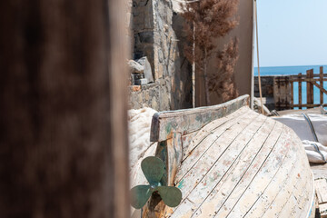 Old weathered wooden boat resting upside down on land, revealing its propeller, with a stone wall and sea visible in background