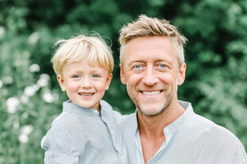 A smiling Caucasian man holds a young boy with blond hair. They are outdoors surrounded by greenery and flowers, enjoying a sunny day together.
