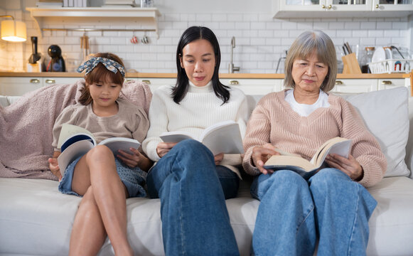 Asian three generations family little girl, mom, and grandma reading books together on the sofa in a cozy living room. Bookworm concept highlighting family bonding, education, and love for reading.