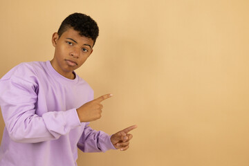Young brown skinned Latino man wearing a lilac sweatshirt pointing with his fingers at a copy space on a light brown background, advertising space to promote