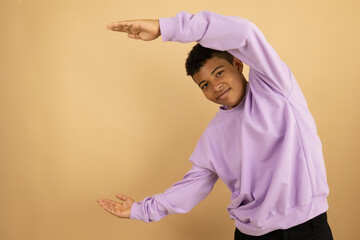 Brown skinned Latino teenager wearing a lilac sweatshirt pointing with both hands at a copy space on a light brown background, advertising space to promote