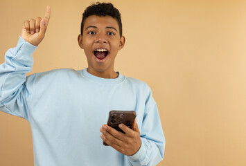 Cheerful young Latino man with brown skin wearing a pastel blue sweatshirt raising his hand with a surprised expression having an idea while holding his cell phone on a light brown background