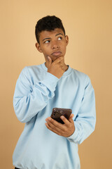 Vertical image of a dark skinned Latino teenager wearing a pastel blue sweatshirt with a thoughtful expression while holding his cell phone on a light brown background with copy space