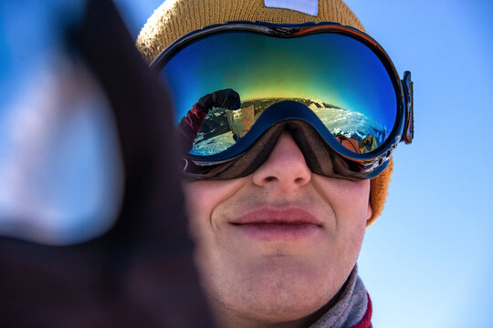 Close-up of smiling man wearing ski goggles with reflection of snowy mountains and camper, winter outdoor adventure