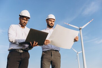 Engineers discussing blueprint and using laptop near wind turbines