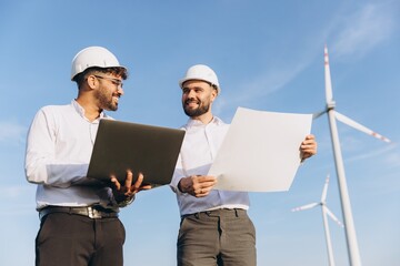 Engineers discussing blueprint and using laptop near wind turbines
