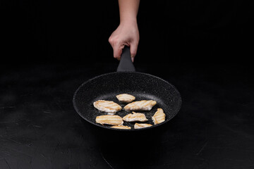 Hand holding frying pan with sizzling chicken fillets cooking on black background, showcasing...
