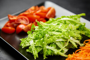 Fresh vegetables including chopped lettuce, sliced tomatoes, and grated carrots arranged on a black plate, showcasing vibrant colors and healthy ingredients for culinary presentation
