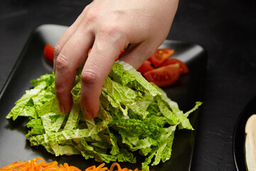 Hand reaching for fresh green lettuce on a black plate, surrounded by vibrant shredded carrots and sliced tomatoes, showcasing healthy food preparation and culinary creativity