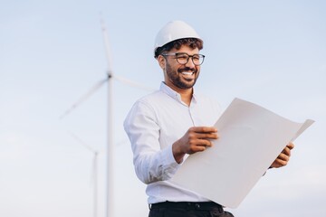 Smiling engineer reading blueprint in front of wind turbines