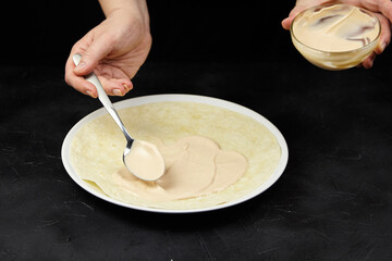 Hands applying creamy sauce onto a thin pancake on a white plate, showcasing culinary skills and preparation techniques in a modern kitchen setting