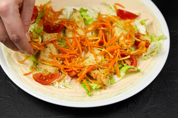 Hand adding fresh vegetables to a tortilla wrap filled with shredded carrots, lettuce, and tomatoes, showcasing vibrant colors and healthy meal preparation