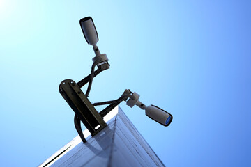 Two black security cameras mounted on a curved metal pole against a clear blue sky. The cameras...