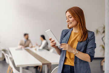 Ginger businesswoman standing at minimalistic corporate office with tablet in hands and smiling at...