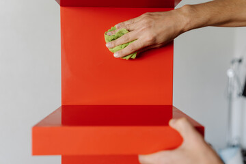 Close-up cropped shot of male hands cleaning red bookshelf with green rag, ensuring spotless and...