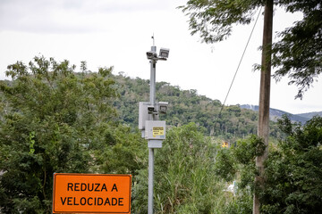 Radar fixo com placa “Reduza a Velocidade” em estrada cercada por vegetação. Imagem sobre segurança no trânsito e controle de velocidade em áreas rurais.