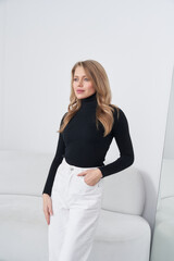 Blonde woman in black turtleneck and white pants, standing near light-colored sofa, one hand in...