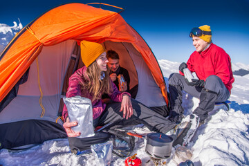 Smiling hikers enjoying winter camping in snowy mountains. Woman holding freeze-dried food pouch outside tent © XArt
