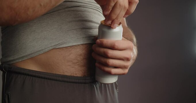 Modern Adult man in sweatpants and a T-shirt with a bare hairy beer belly opens a metal can with a drink. Close-up on the stomach and hands