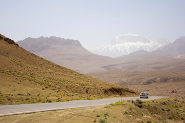 A car winds along a remote road through Yazd’s arid terrain, with dramatic snow-capped mountains rising in the background beneath a sunlit sky.