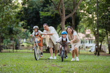 A family of four riding bikes in a park. A man is helping a child on a bike
