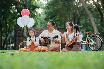A family of four is sitting on the grass in a park. A man is playing a guitar and the children are...