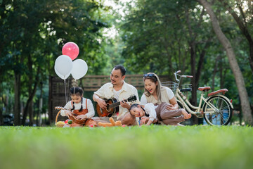 A family of four is sitting on the grass in a park. The father is playing the guitar while the...