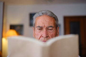 Close-up of elderly man reading a book at home with face partially hidden.