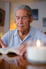 Vertical view of an elderly man reading a book with a candle lit in front of him at home.