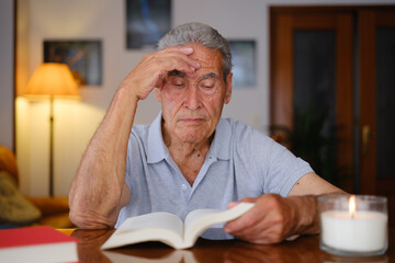 Elderly man reading at home, touching his forehead in a moment of mental fatigue, sitting beside a lit candle at a wooden table.