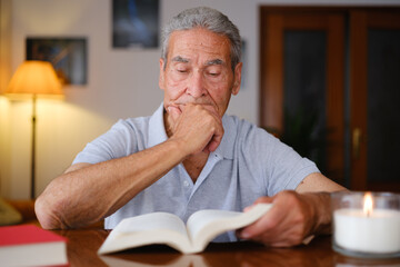 Elderly man reading a book at home, resting his chin on his hand with a focused and introspective expression, beside a lit candle.