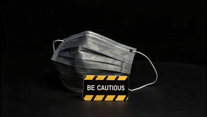 Naklejka premium A protective face mask is placed beside a sign that reads be cautious against a dark background, urging vigilance