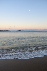 Picturesque sunset on a sandy beach in Skiathos, Greece. Gentle pastel colours glow in the sky above calm beach waves, backdropped by Greek island landscapes.