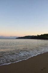 Picturesque sunset on a sandy beach in Skiathos, Greece. Gentle pastel colours glow in the sky above calm beach waves, backdropped by Greek island landscapes.
