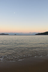 Picturesque sunset on a sandy beach in Skiathos, Greece. Gentle pastel colours glow in the sky above calm beach waves, backdropped by Greek island landscapes.