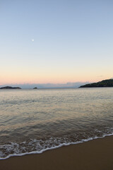 Picturesque sunset on a sandy beach in Skiathos, Greece. Gentle pastel colours glow in the sky above calm beach waves, backdropped by Greek island landscapes.
