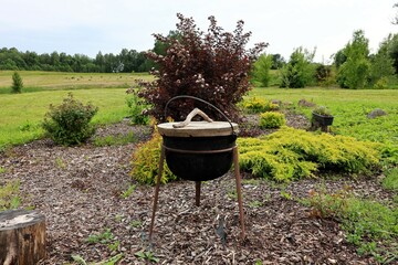 Old-fashioned black cast iron cauldron with a wooden lid standing on tripod legs, placed in a...