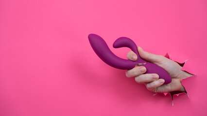 Female hand sticking out with raspberry sex toy from pink paper background.
