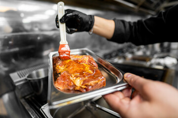 Chef basting raw meat with marinade using a brush in a professional kitchen. Close-up of gloved...