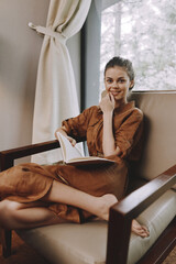 Cozy relaxation of a young woman in a brown dress, sitting comfortably in a chair, reading a book,...