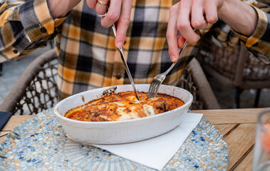 Person cutting baked pasta Bolognese with fork and knife at outdoor restaurant