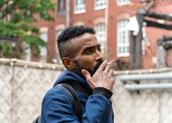Young handsome African American man smoking cigarette in alley. Lips blowing smoke on blurred background. Portrait gangster smokes marijuana or cannabis. Portrait of a black guy smoking a cigarette.
