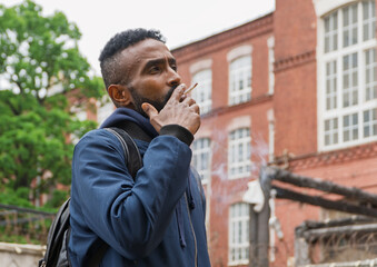 Young handsome African American man smoking cigarette in alley. Lips blowing smoke on blurred background. Portrait gangster smokes marijuana or cannabis. Portrait of a black guy smoking a cigarette.