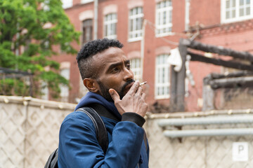 Young handsome African American man smoking cigarette in alley. Lips blowing smoke on blurred background. Portrait gangster smokes marijuana or cannabis. Portrait of a black guy smoking a cigarette.
