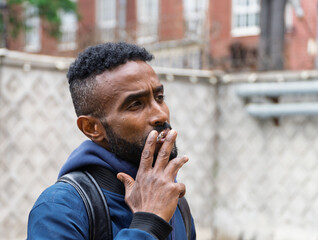 Young handsome African American man smoking cigarette in alley. Lips blowing smoke on blurred background. Portrait gangster smokes marijuana or cannabis. Portrait of a black guy smoking a cigarette.