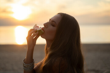 A woman in a green palm-print bikini kneels on a white blanket on the sand, enjoying a slice of...