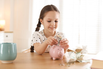 Girl putting pocket money into piggybank at table indoors