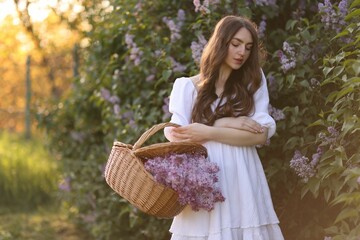 Beautiful woman with basket of lilac flowers near bush outdoors in morning