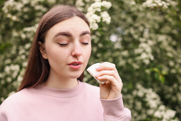 Woman with paper tissue suffering from seasonal pollen allergy near blossoming shrub on spring day,...